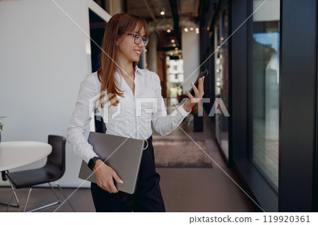 A Young Professional Woman is working in an Office with both a Laptop and a Smartphone in hand 119920361