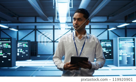System administrator walks in data center used for managing gear energy consumption, working on tablet. Close up of IT specialist using device in workspace used for automation in server room, camera B 119920901