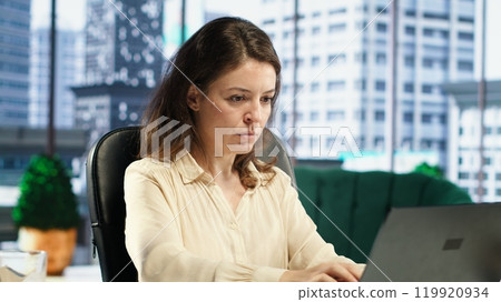 Stressed overwhelmed business leader working in discomfort with a headache, feeling overburdened and uncomfortable at the office. Female executive deals with anxiety and fatigue. Camera A. 119920934