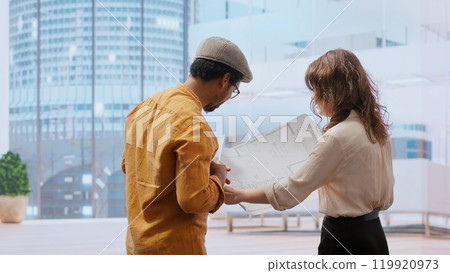 Business professional and real estate broker reviewing blueprints on laptop for high class office leasing contract, discussing the architectural design and layout. Planning to buy property. Camera B. 119920973