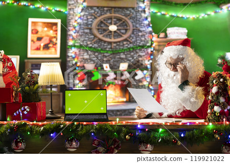 Father Christmas reading letters from children next to green screen display in his cabin at the North Pole, spreading holiday tradition during winter festivity. Finding gift wishes. Father Christmas reading letters from children next to green screen display in his cabin at the North Pole, spreading holiday tradition during winter festivity. Finding gift wishes. 119921022