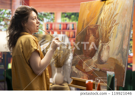 Skilled artist stands before an easel painting a watercolor rustic scene. Woman showing her talent and creativity as she mixes dyes and pigments, producing a multicolored masterpiece. Skilled artist stands before an easel painting a watercolor rustic scene. Woman showing her talent and creativity as she mixes dyes and pigments, producing a multicolored masterpiece. 119921046