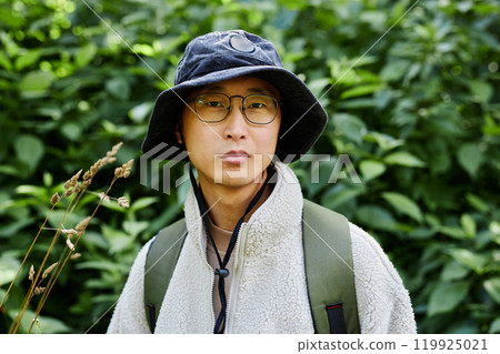 Vibrant portrait of young Asian man as male tourist looking at camera against forest greenery and wearing eyeglasses copy space 119925021