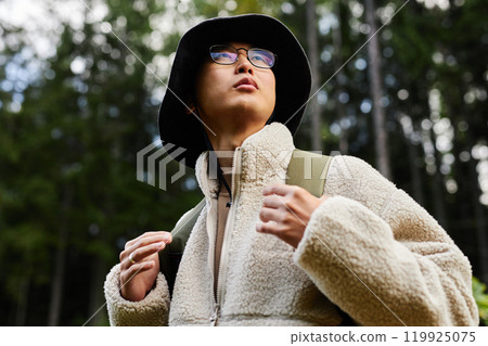 Portrait of young Asian man backpacking in forest and looking up with focus on eyeglasses and comfy sherpa coat copy space 119925075