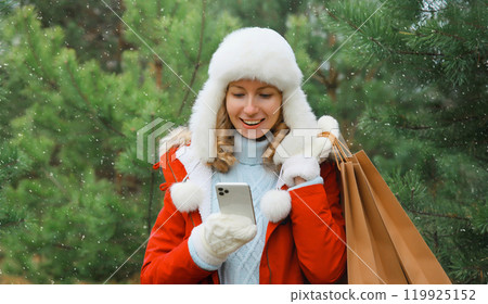 Happy smiling woman with phone, shopping bag in winter against Christmas tree, girl using smartphone Happy smiling woman with phone, shopping bag in winter against Christmas tree, girl using smartphone 119925152