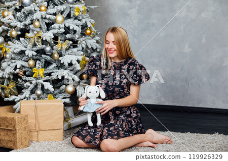 Blonde woman in dress smiling with gifts at Christmas tree for new year 119926329