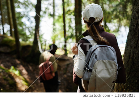 Back view of young woman with backpack walking along forest path while travelling with group of friends in nature, copy space 119926825