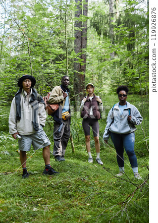Multiethnic group of tourists standing outdoors on forest path and looking at camera while hiking together 119926876