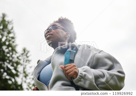 Dramatic low angle portrait of African American young woman as female tourist with backpack wearing sunglasses and walking outdoors against sky copy space 119926992