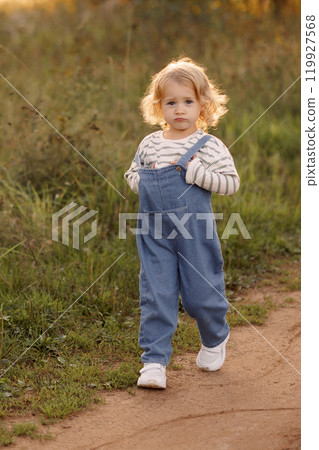 A young blonde girl in a blue jumpsuit walks along a dirt road in the park A young blonde girl in a blue jumpsuit walks along a dirt road in the park 119927568