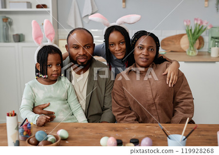Family enjoying Easter celebration wearing bunny ears while painting eggs in kitchen setting with flowers in background Family enjoying Easter celebration wearing bunny ears while painting eggs in kitchen setting with flowers in background 119928268