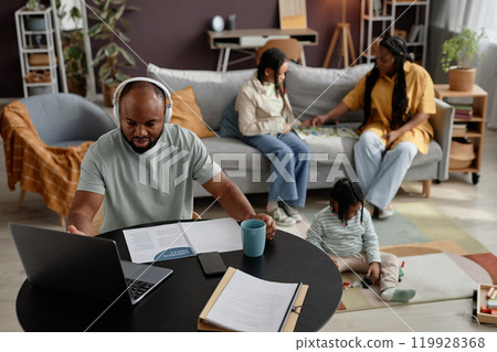African American father engaging in work with laptop and headphones while family interacting in background creating a warm family workspace environment African American father engaging in work with laptop and headphones while family interacting in background creating a warm family workspace environment 119928368