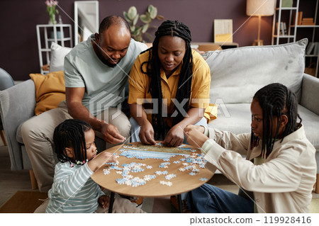 Family gathered around wooden table solving puzzle, focusing on pieces and working together. Living space is warm and inviting, with plants and lamps adding ambiance Family gathered around wooden table solving puzzle, focusing on pieces and working together. Living space is warm and inviting, with plants and lamps adding ambiance 119928416