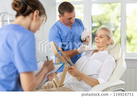 Man dermatologist examines skin of senior womans face, female nurse records data in client card Man dermatologist examines skin of senior womans face, female nurse records data in client card 119928731