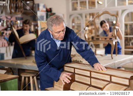 Ederly craftsman polishes tabletop of old chest of drawers with rounded drawers with soft cloth 119928817