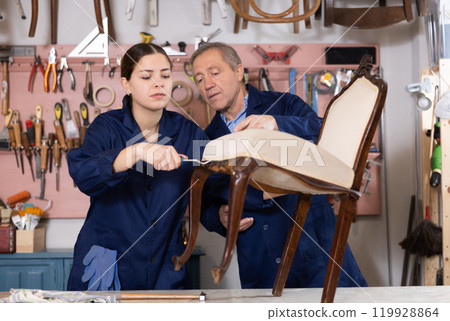 Professional mature craftsman restorer in uniform helping young craftswoman renewing antique chair in repair shop Professional mature craftsman restorer in uniform helping young craftswoman renewing antique chair in repair shop 119928864