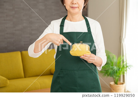 A senior woman housewife/housekeeper wearing a green apron standing in the living room at home pointing at her purse A senior woman housewife/housekeeper wearing a green apron standing in the living room at home pointing at her purse 119929050