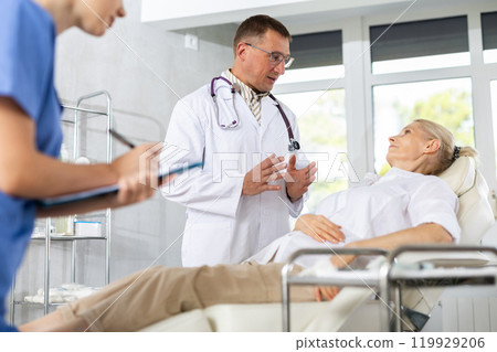 Male beautician, together with a nurse, examines the face of an mature woman and plans facelift procedures. 119929206