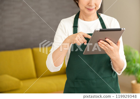 A senior woman housewife/housekeeper wearing a green apron smiling while looking at a tablet in the living room at home 119929348