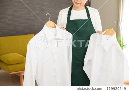 A senior female housewife/housekeeper wearing a green apron holding a Y-shirt in the living room at home A senior female housewife/housekeeper wearing a green apron holding a Y-shirt in the living room at home 119929349