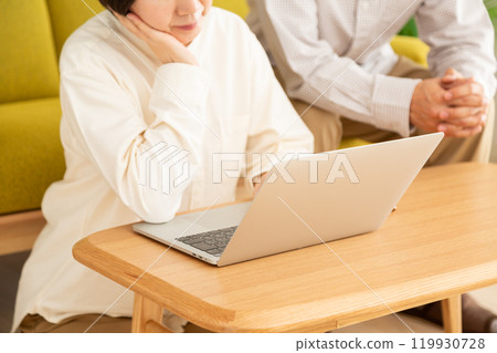 A middle-aged to senior couple, a man and a woman, looking at a computer in the living room and worrying A middle-aged to senior couple, a man and a woman, looking at a computer in the living room and worrying 119930728