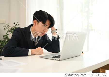 Young man in a suit sitting at a desk, frustrated while working on the laptop 119931127