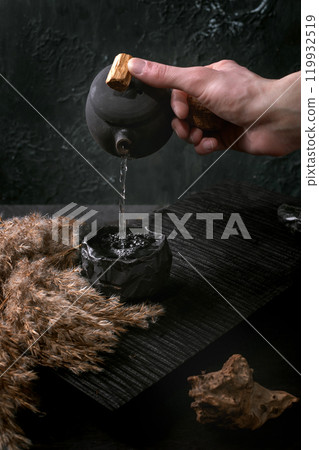 Chinese Tea Ceremony. A man's hand pours tea from a traditional handmade ceramic black teapot with wooden handle into a cup on a dark tea table. Autumn Vibe. Dried flowers reed nearby. Chinese Tea Ceremony. A man's hand pours tea from a traditional handmade ceramic black teapot with wooden handle into a cup on a dark tea table. Autumn Vibe. Dried flowers reed nearby. 119932519