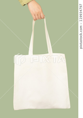Blank white tote bag canvas fabric with handle mock up design. Close up of woman hand holding eco or reusable shopping bag on green background. No plastic bag and ecology concept. Vertical. 119934797