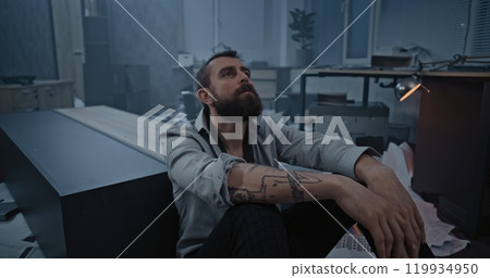 Dolly shot of frustrated office worker sitting on the floor in destroyed office room 119934950