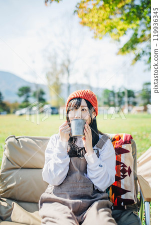 Young woman wearing knitted hat relaxing with a drink in camp 119936345