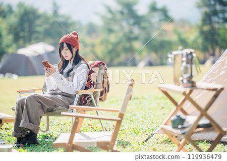 一位戴著針織帽的年輕女子坐在營地的椅子上操作智慧型手機 119936479
