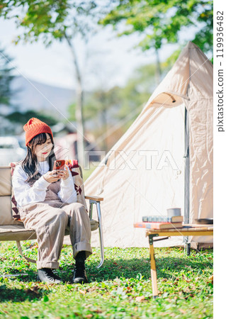 A young woman in a knitted hat operating a smartphone while sitting on a chair at a campsite A young woman in a knitted hat operating a smartphone while sitting on a chair at a campsite 119936482