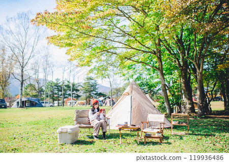 A young woman in a knitted hat operating a smartphone while sitting on a chair at a campsite A young woman in a knitted hat operating a smartphone while sitting on a chair at a campsite 119936486