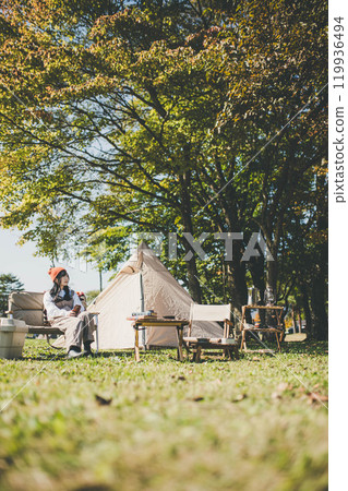 A young woman in a knitted hat operating a smartphone while sitting on a chair at a campsite 119936494