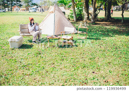 一位戴著針織帽的年輕女子坐在營地的椅子上操作智慧型手機 119936499