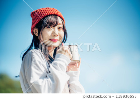 A young woman wearing a knitted hat drinking a drink under a blue sky A young woman wearing a knitted hat drinking a drink under a blue sky 119936653