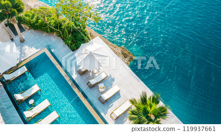 Aerial view of luxurious poolside with sun loungers, umbrellas, and lush greenery overlooking the serene turquoise waters in a tropical paradise. Aerial view of luxurious poolside with sun loungers, umbrellas, and lush greenery overlooking the serene turquoise waters in a tropical paradise. 119937661
