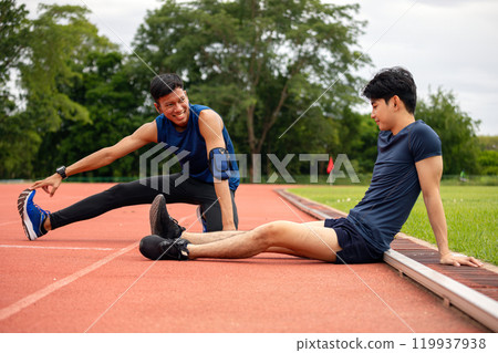 Two male athletes stretching and preparing for training on track, with one athlete reaching for his foot and smiling Two male athletes stretching and preparing for training on track, with one athlete reaching for his foot and smiling 119937938