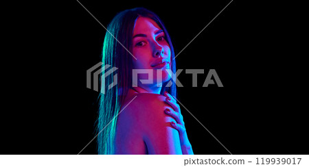 Portrait of young attractive woman posing in bright neon light, gently touching her shoulder against black studio background. 119939017