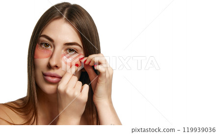 Woman with bare shoulders applying under-eye hydrogel patches for hydration against white background. With copy space. Woman with bare shoulders applying under-eye hydrogel patches for hydration against white background. With copy space. 119939036