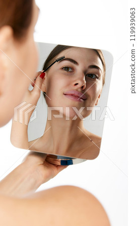 Woman brushing her eyebrows while looking in mirror, highlighting precise grooming against white background. Woman brushing her eyebrows while looking in mirror, highlighting precise grooming against white background. 119939063