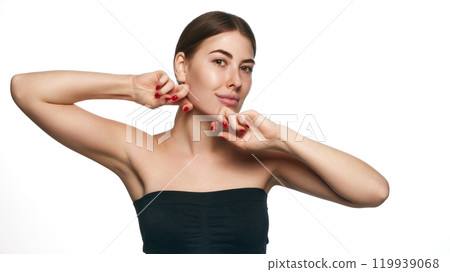 Portrait of young woman performing facial exercise by gently pinching her cheeks, chin against white background. 119939068