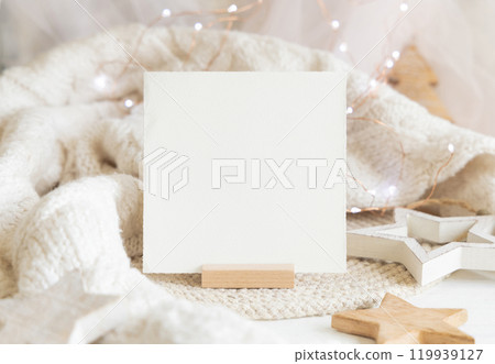 Square paper card near white sweater and wooden Christmas decor and lights, mockup 119939127
