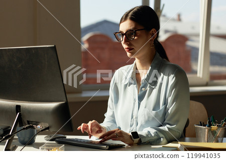 Portrait of successful young woman wearing glasses and using computer by window lit by sunlight, copy space 119941035
