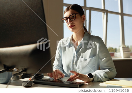 Portrait of successful young businesswoman using computer enjoying workday in office lit by sunlight, copy space 119941036