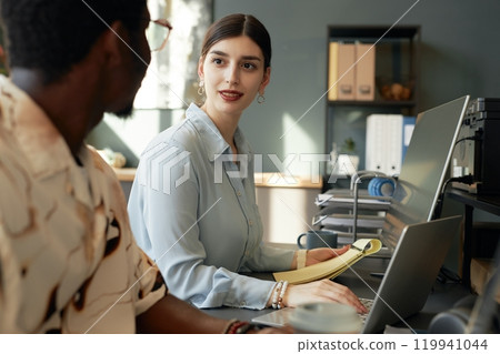 Two people working together in office, focus on smiling young woman talking to colleague while using computer 119941044