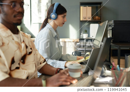 Side view portrait of two people using computers in row in office, focus on young woman wearing headphones and typing, copy space 119941045