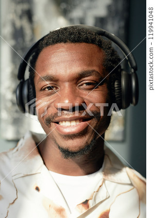 Vertical closeup portrait of African American man wearing big headphones and smiling happily at camera lit by bright sunlight 119941048