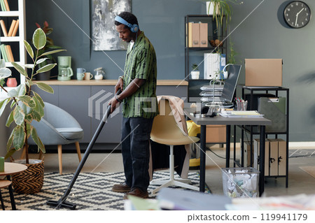 Side view full length portrait of young African American man vacuuming carpet and listening to music while cleaning floors in office, copy space Side view full length portrait of young African American man vacuuming carpet and listening to music while cleaning floors in office, copy space 119941179