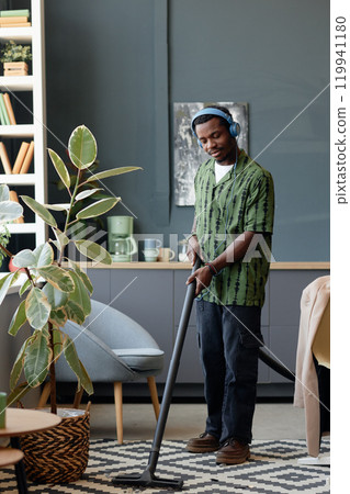 Vertical full length portrait of young African American man wearing headphones while vacuuming carpet and cleaning floors in office Vertical full length portrait of young African American man wearing headphones while vacuuming carpet and cleaning floors in office 119941180